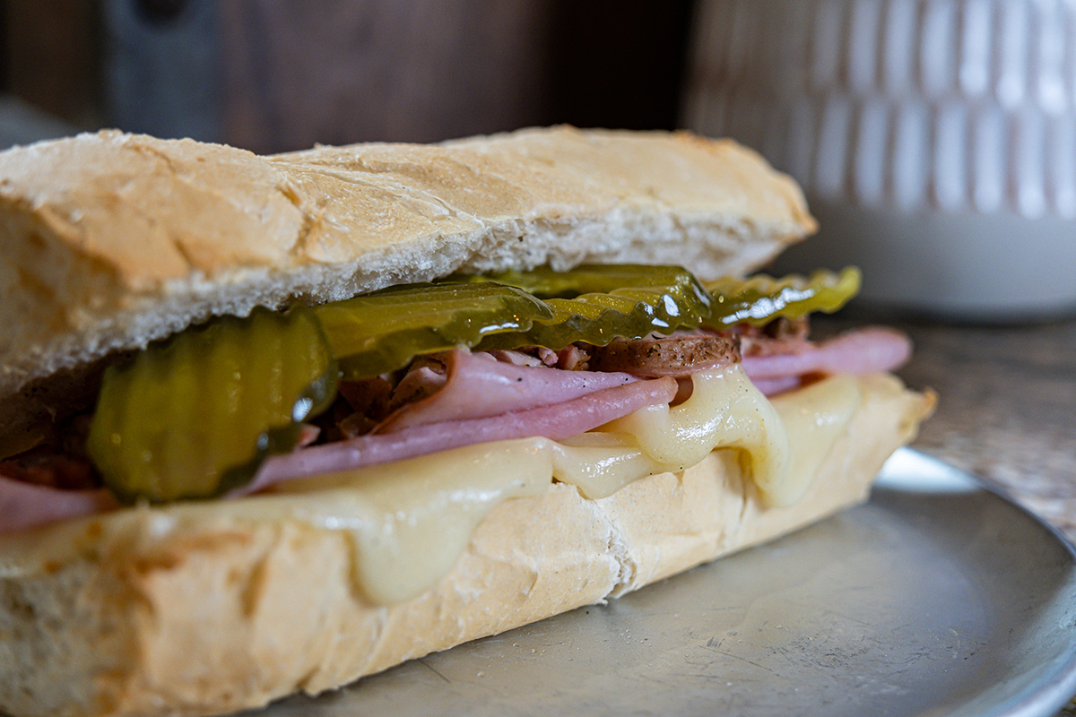 Cuban sandwich on a metal plate, featuring sliced ham, melted cheese, pickles, and crusty bread.