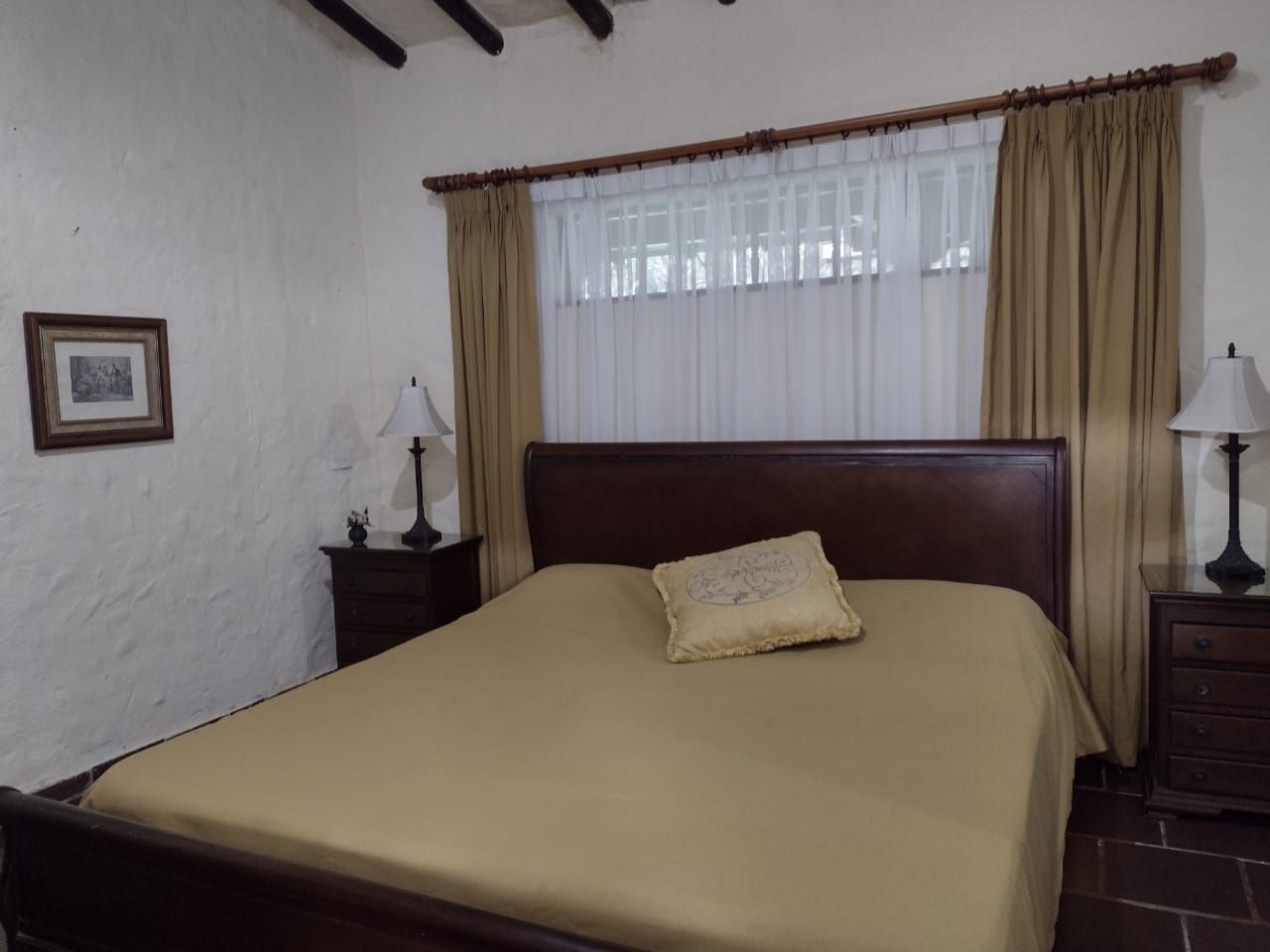 Dormitorio con cama de madera oscura, mesitas de noche, ropa de cama y cortinas color beige, y obras de arte en la pared.
