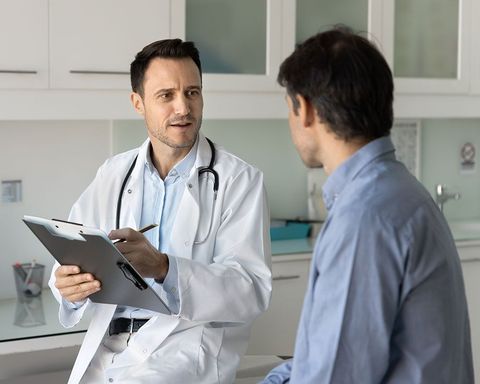 A doctor in a white coat with a stethoscope around their neck writes on a clipboard while speaking to a seated patient.