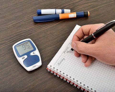 A hand writes in a notebook beside a glucose meter reading 95 mg/dL and two insulin pens on a dark wooden surface.