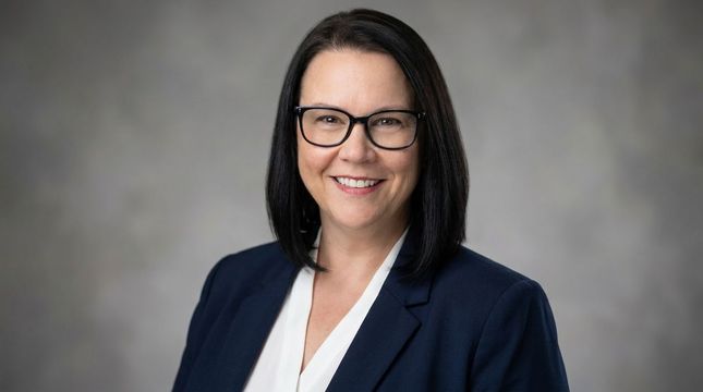 Portrait of a smiling person in glasses, wearing a navy blazer and white blouse against a gray background