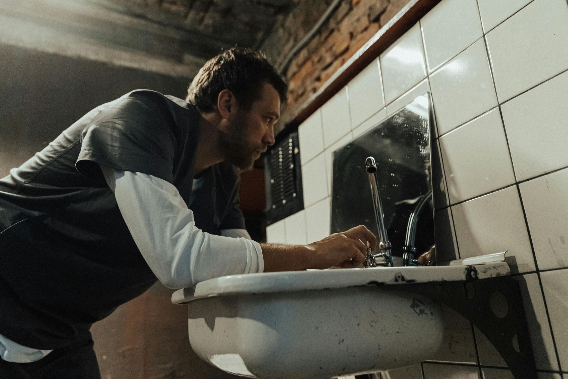Man repairing a bathroom sink under a wall mirror in a tiled room