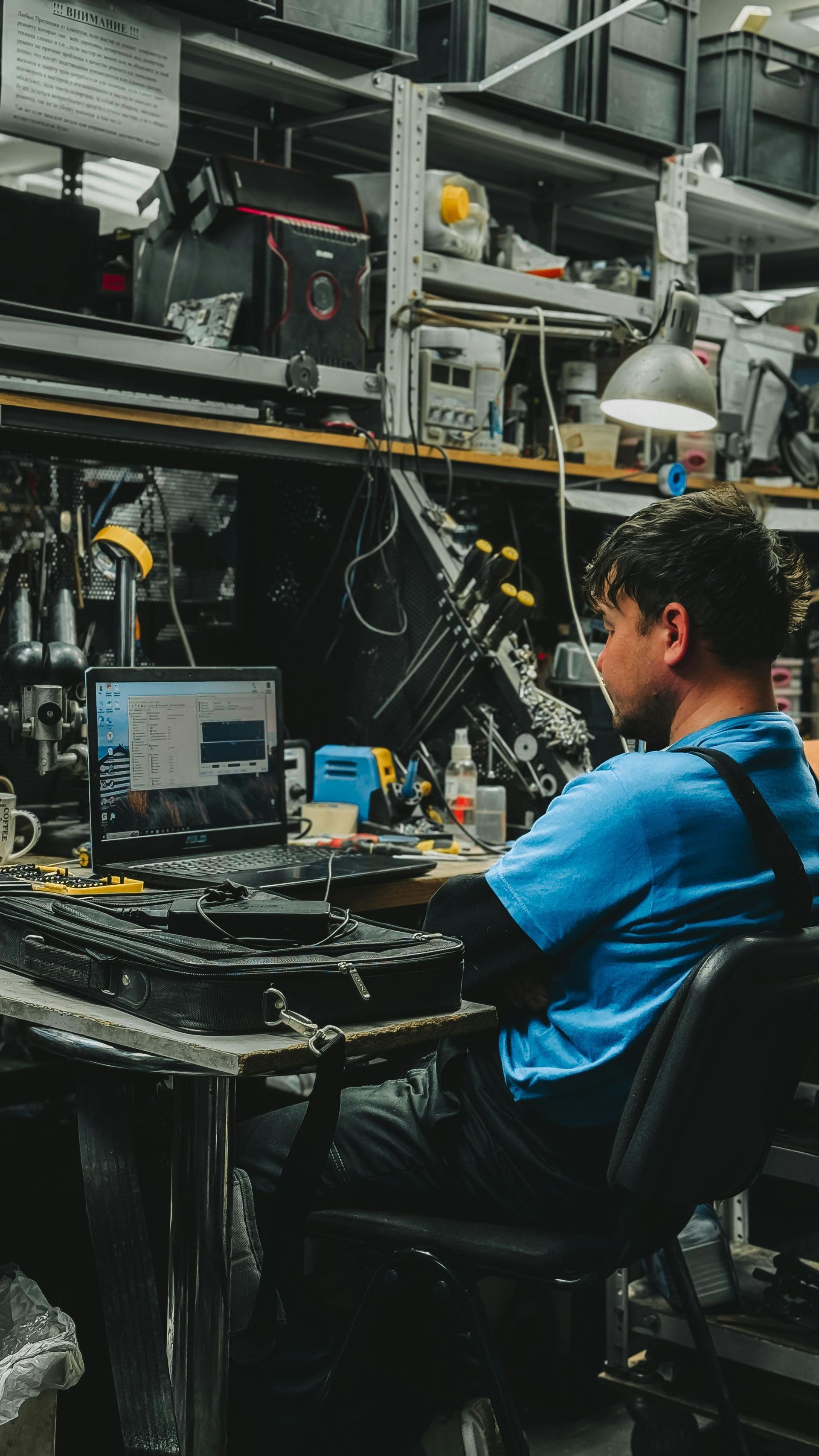 Person seated at a workstation in a cluttered industrial lab, monitoring a computer with cables and machinery nearby