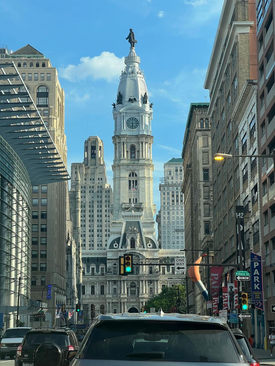 City street view with cars, flanked by tall buildings and a white historic tower in the center under a blue sky
