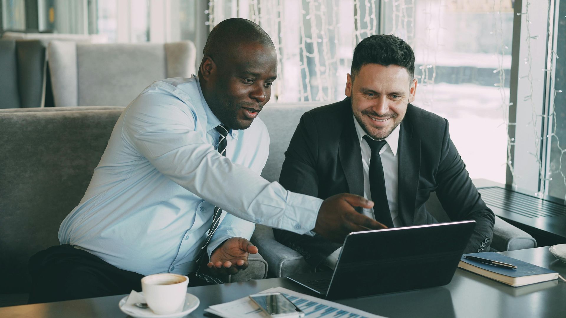 Two coworkers in business attire smiling and reviewing a laptop in a bright office with coffee on the table