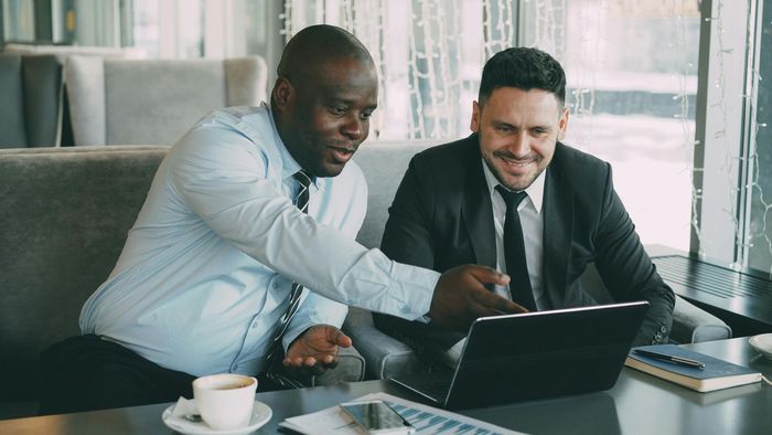 Two coworkers in business attire smiling and reviewing a laptop in a bright office with coffee on the table