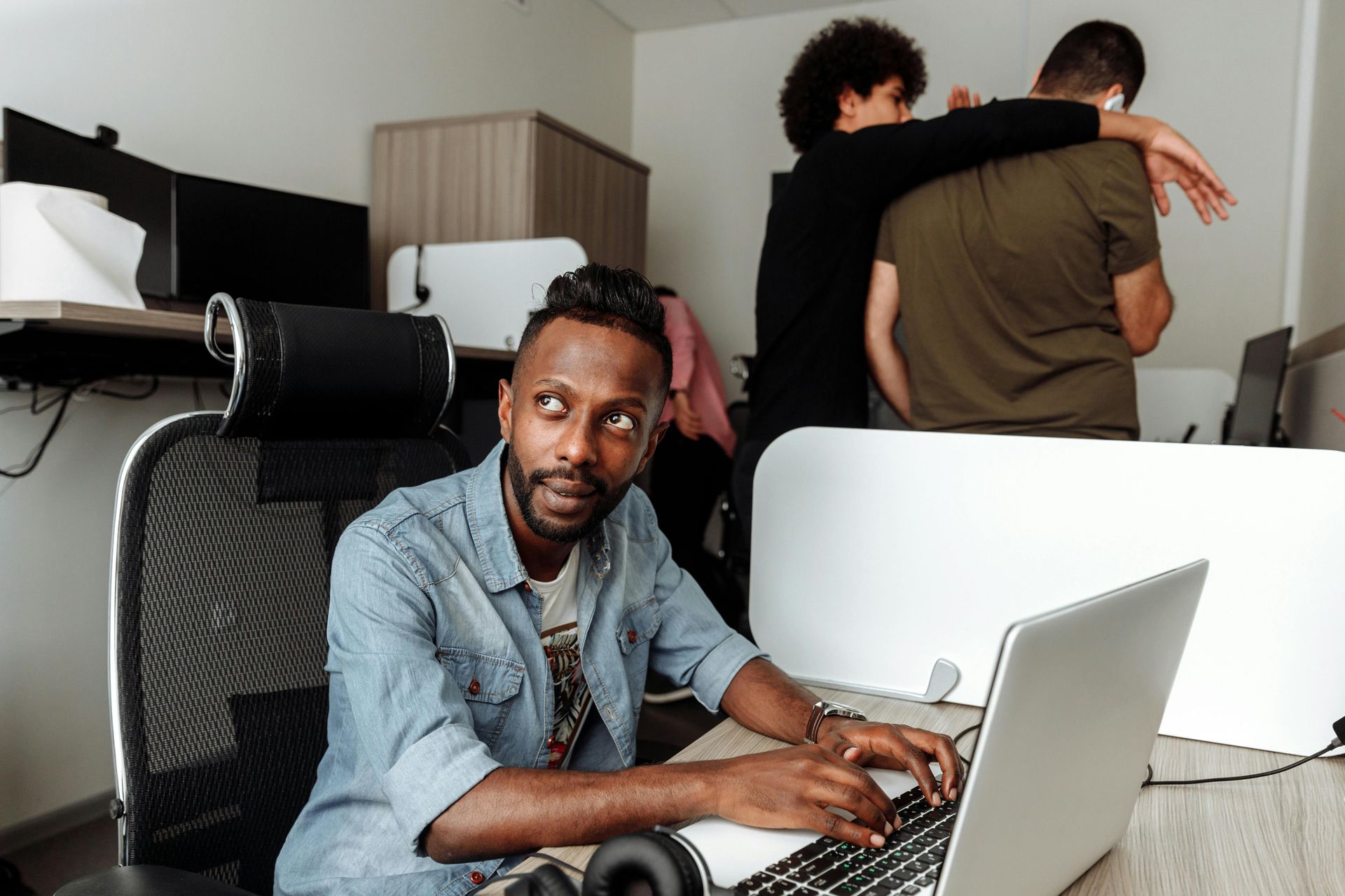 Person using a laptop in a bright office while two people stand together in the background