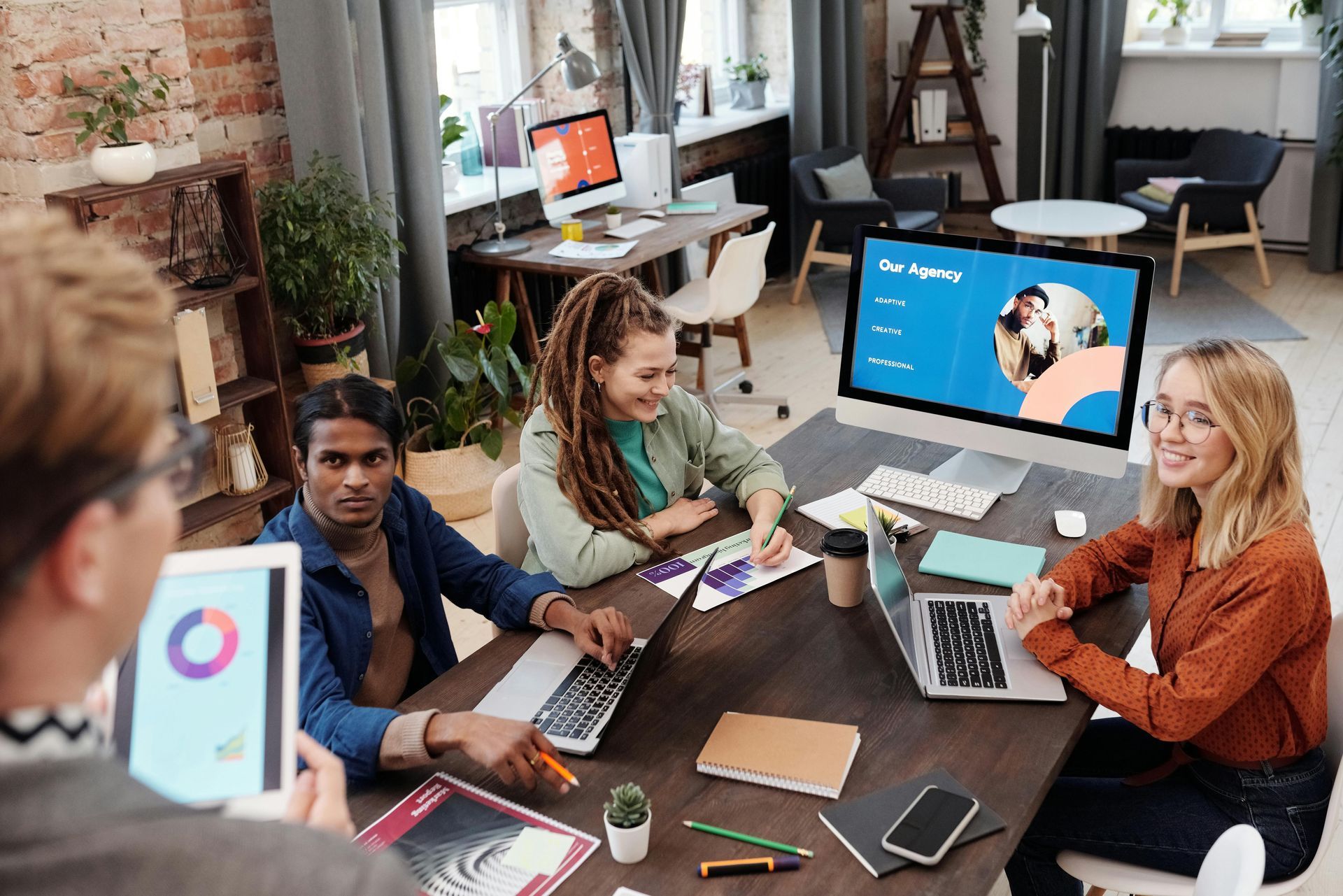 People collaborating around a table in a bright office, using laptops and tablets with a presentation on screen