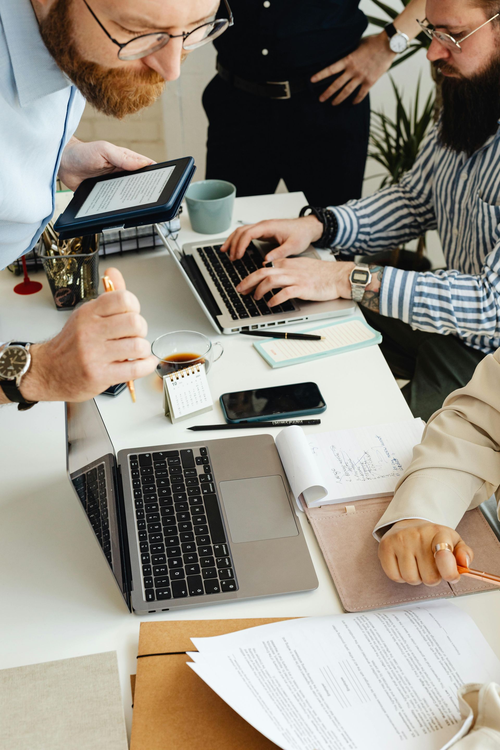 People collaborating at a white table with laptops, tablet, phones, and documents in an office meeting