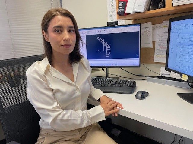 Woman in white shirt sits at a desk, looking at the camera. Computer screens display diagrams and text.