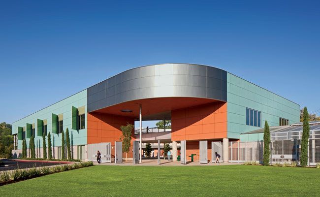 Modern building with green, orange, and silver facade under a clear blue sky, featuring a green lawn in the foreground.