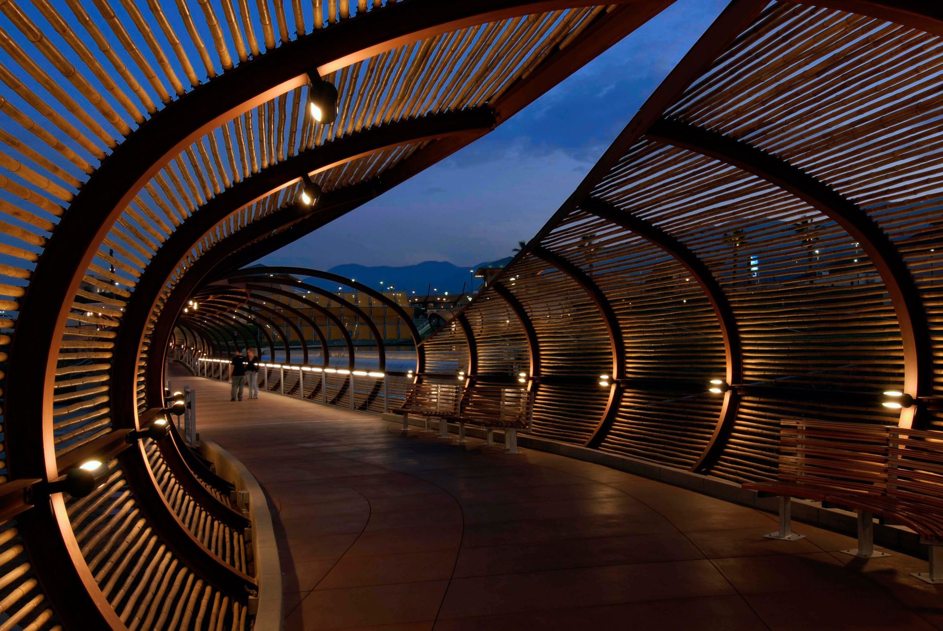A curved walkway tunnel with wooden slats, lit by small lights, against a dusky sky.