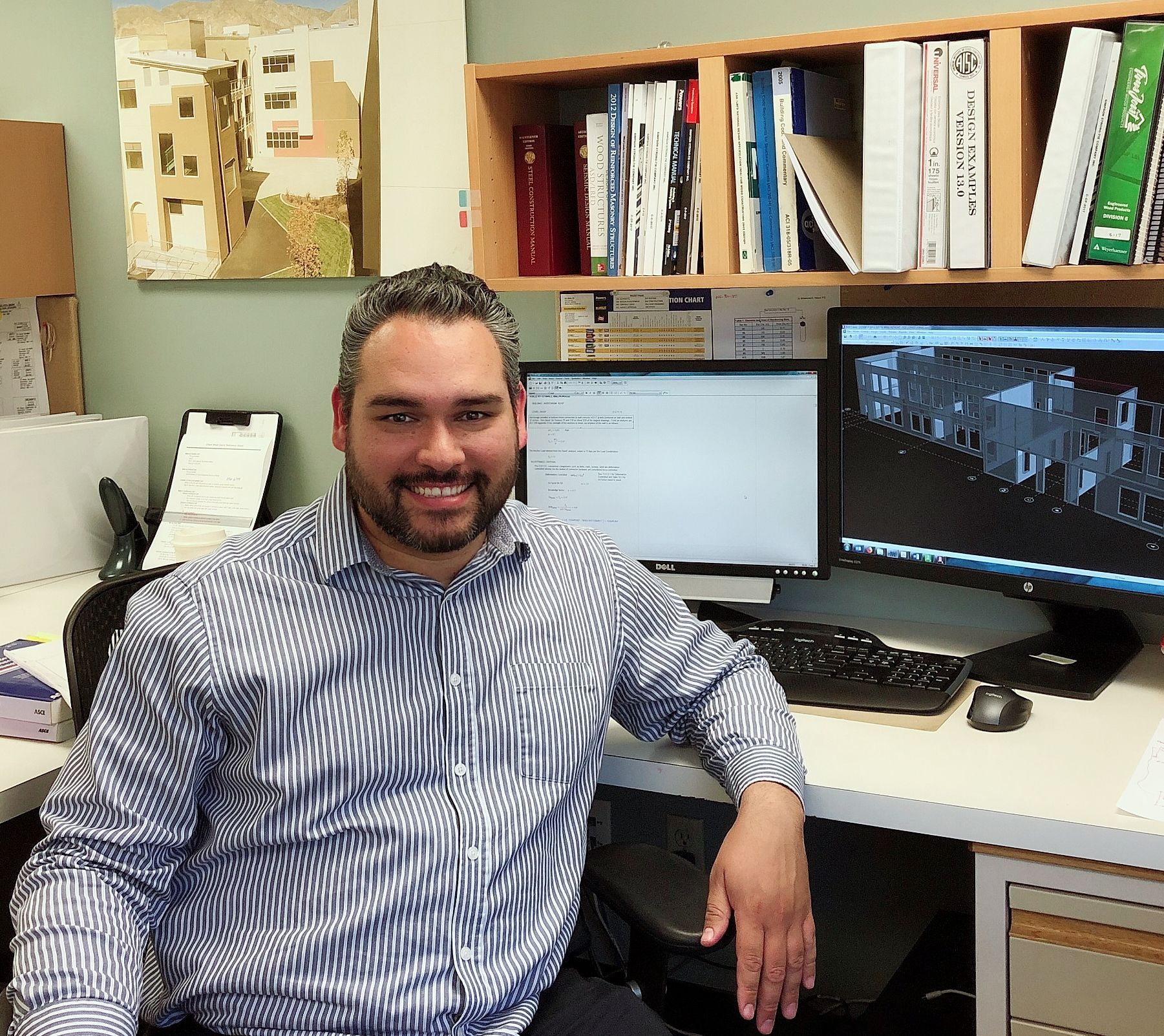 Man in blue striped shirt smiles at his desk with two monitors and bookshelves.