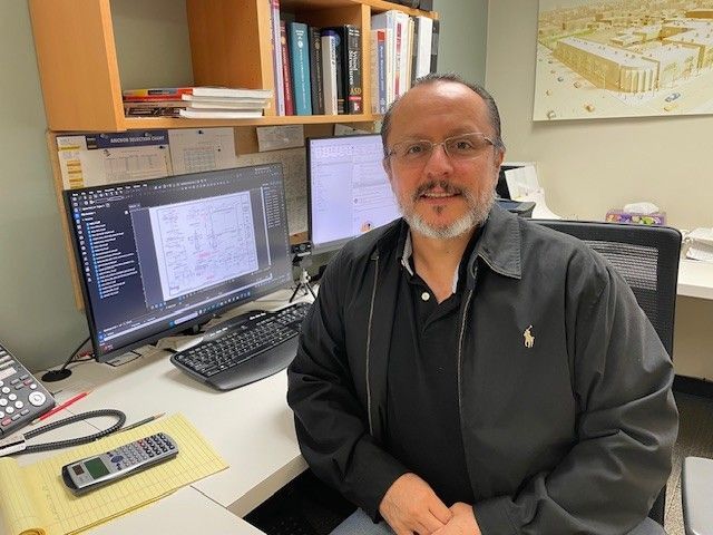 Man with glasses sits at desk in office, two computer monitors, books, black jacket.