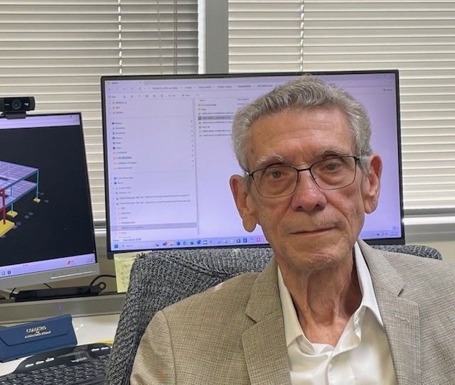 Man with glasses in front of computer monitors, possibly an office setting.
