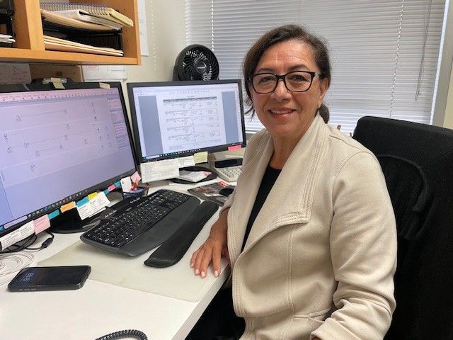 Woman sitting at a desk with two monitors, smiling. She wears glasses, a beige jacket, and a black shirt. Office setting.