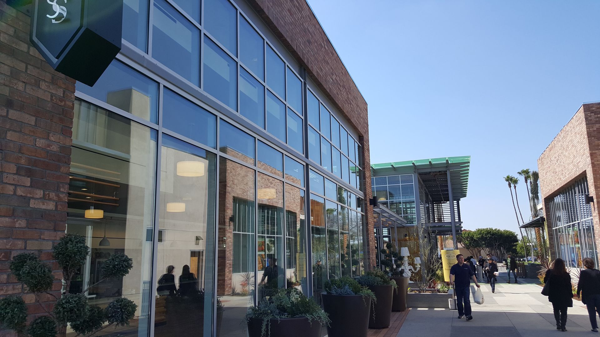 Exterior view of brick building with large glass windows and people walking on a pathway on a sunny day.