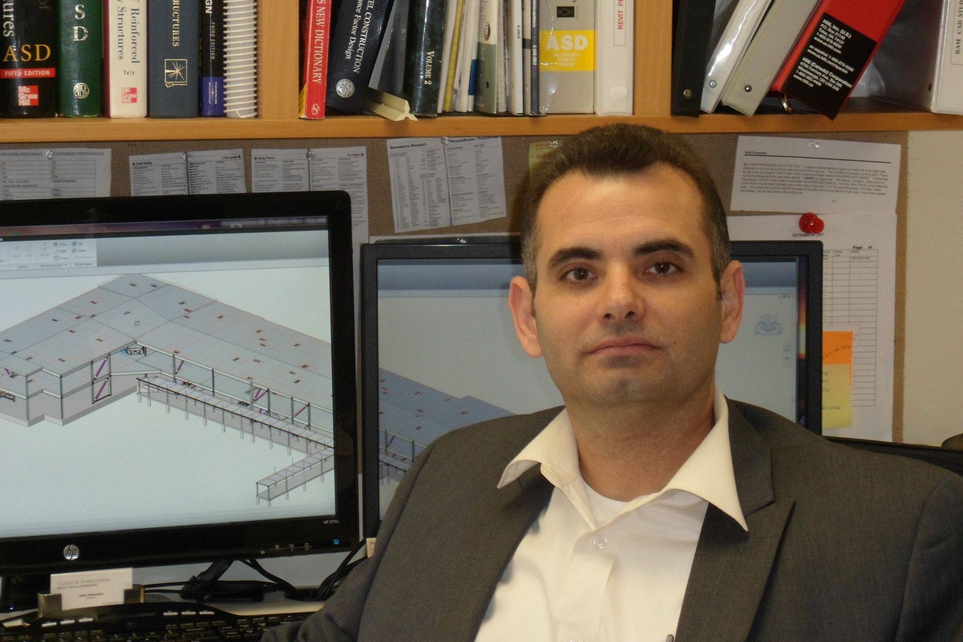 Man in suit sits at computer, a building blueprint displayed on screen, office setting.