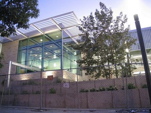 Modern building with glass walls, topped by a metal grid roof, behind a concrete wall and chain link fence.