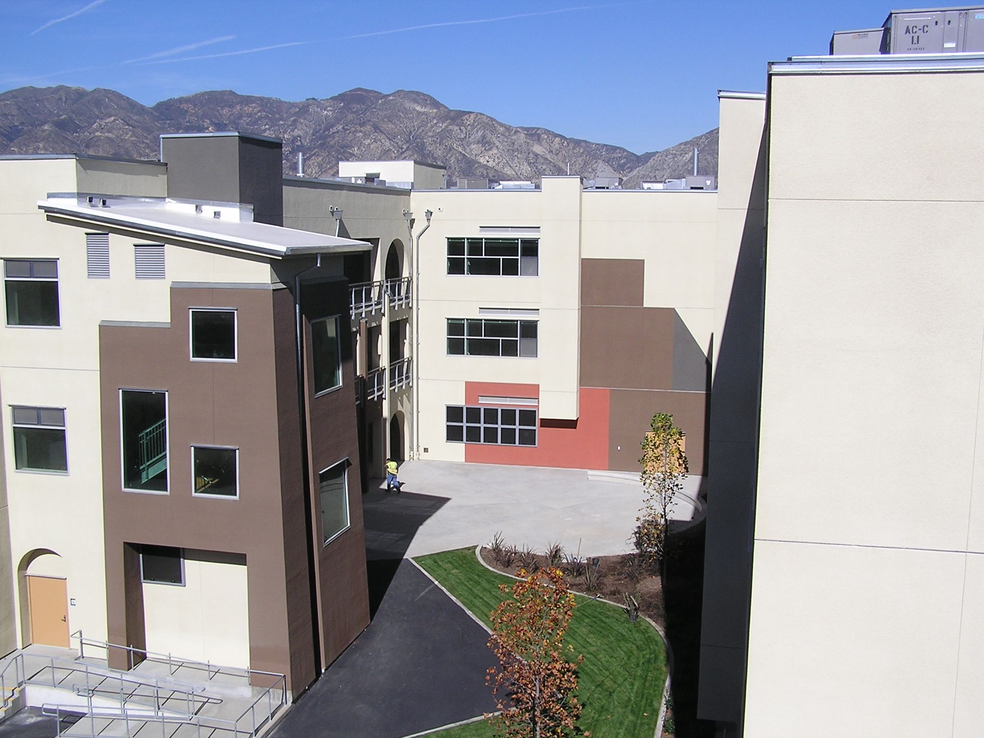 Modern apartment complex, with brown and cream-colored buildings. Green grass, blue sky, and mountains in the background.