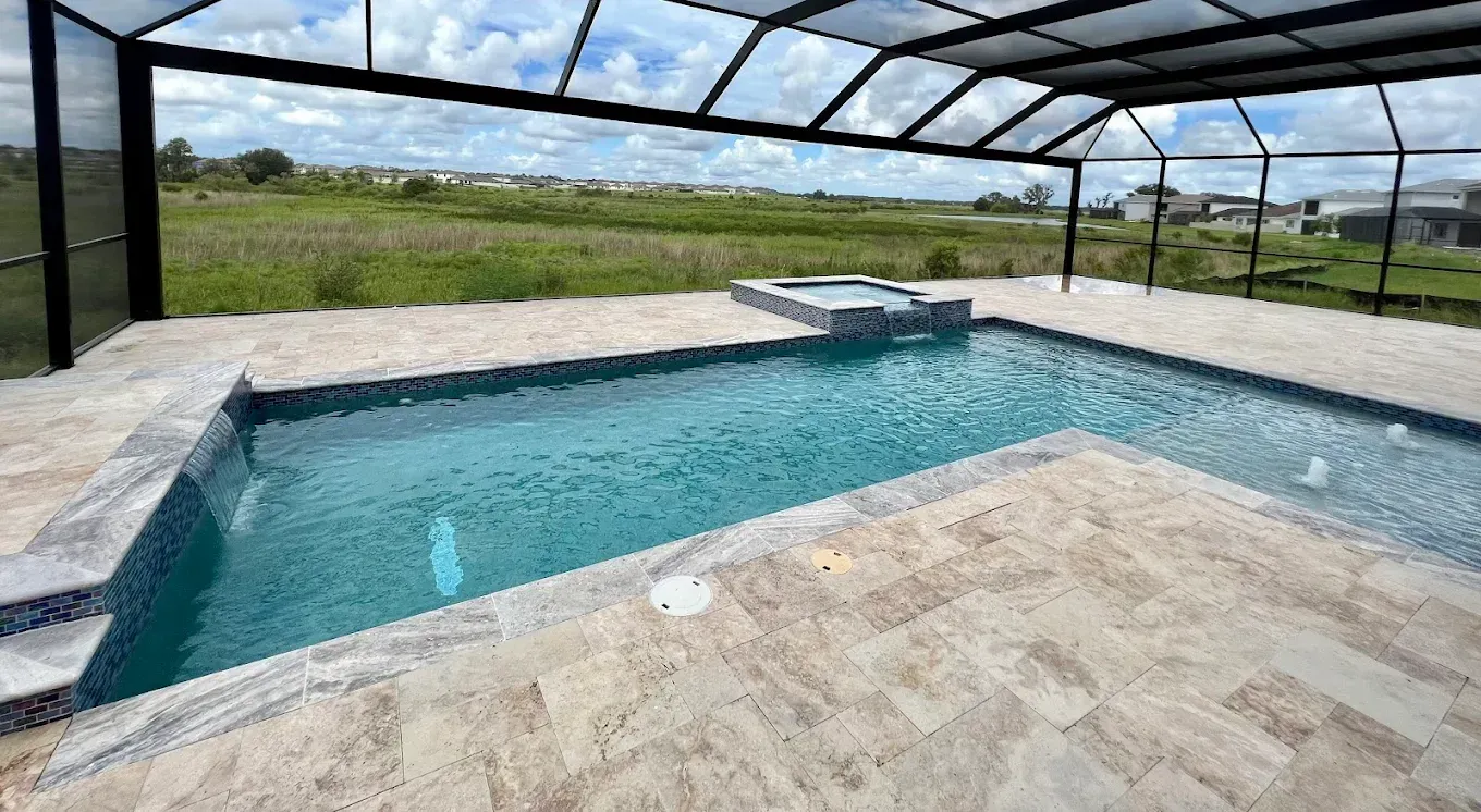 Pool with a spa under a glass roof, surrounded by stone patio, overlooking a field.
