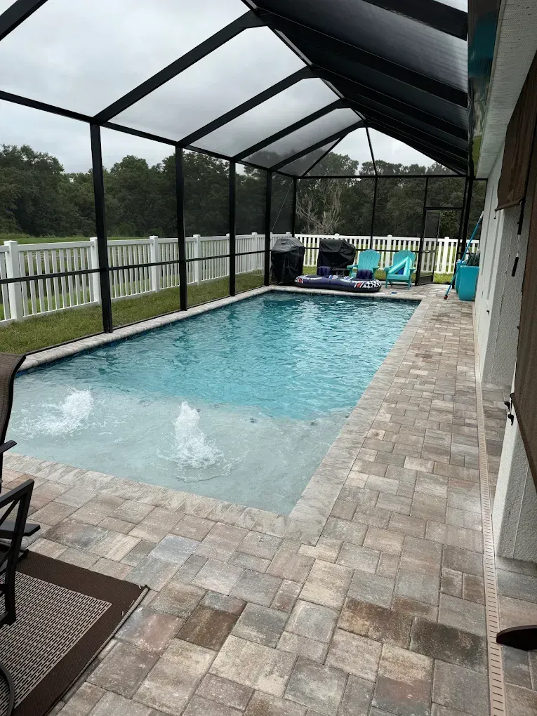 Pool with blue water and fountains, surrounded by pavers, a screened enclosure, and a white fence.