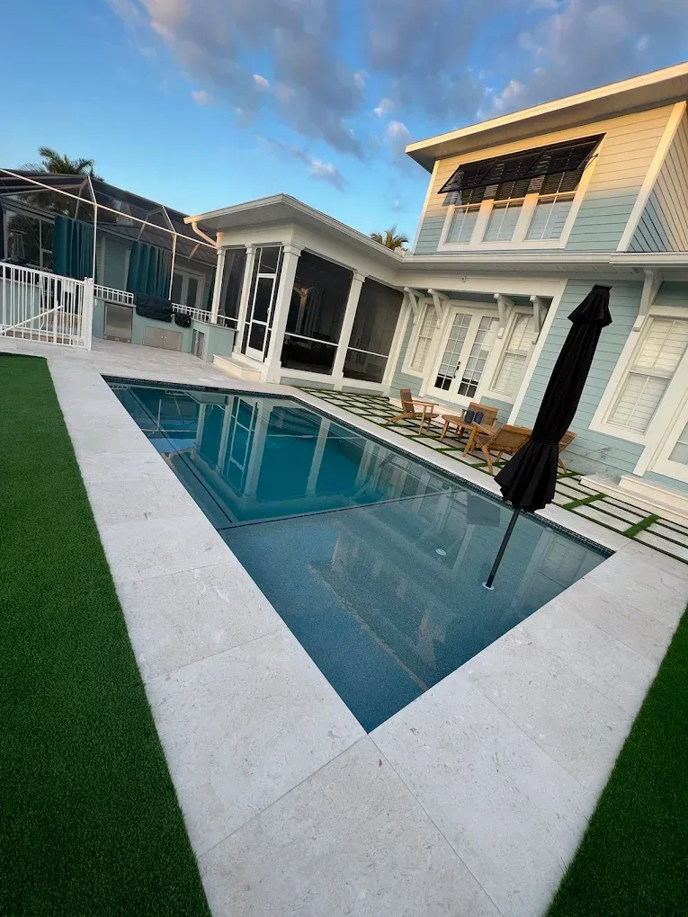 Pool with blue water and white stone patio, next to a light blue house. Green grass surrounds the pool.