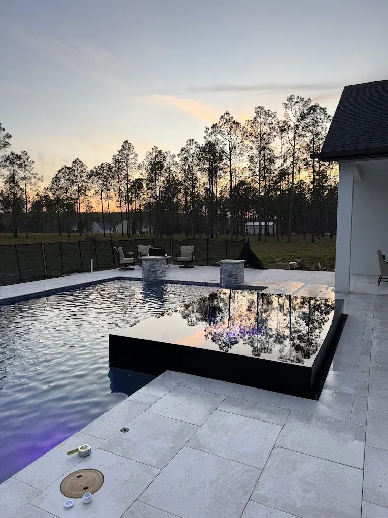Pool with a dark, raised platform reflecting the sky at dusk, surrounded by trees and a modern home.