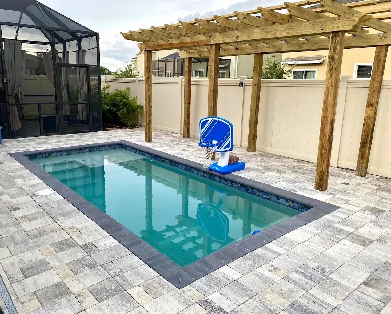 Rectangular pool with basketball hoop, pergola, and stone patio. Beige fence surrounds.