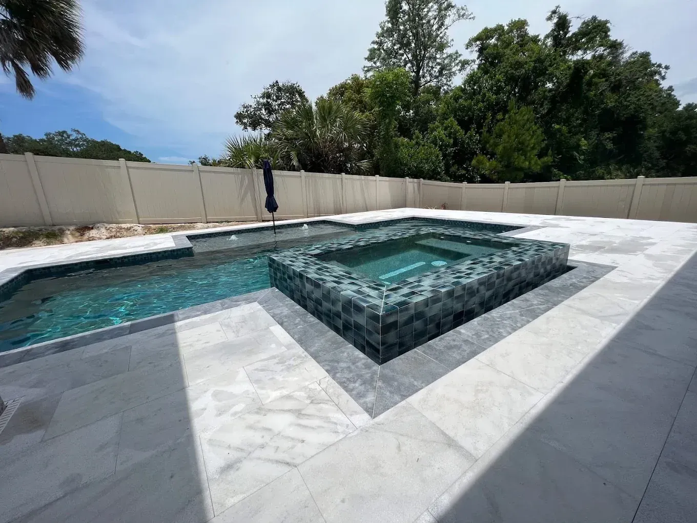 Backyard pool with spa; turquoise water, tiled, light gray patio, white fence, trees, sunny day.