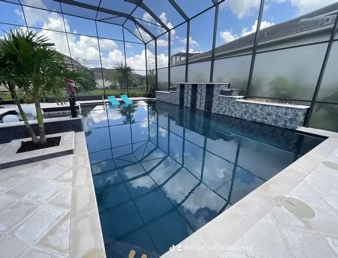 Swimming pool with screen enclosure, stone patio, waterfall feature, and sky reflection.