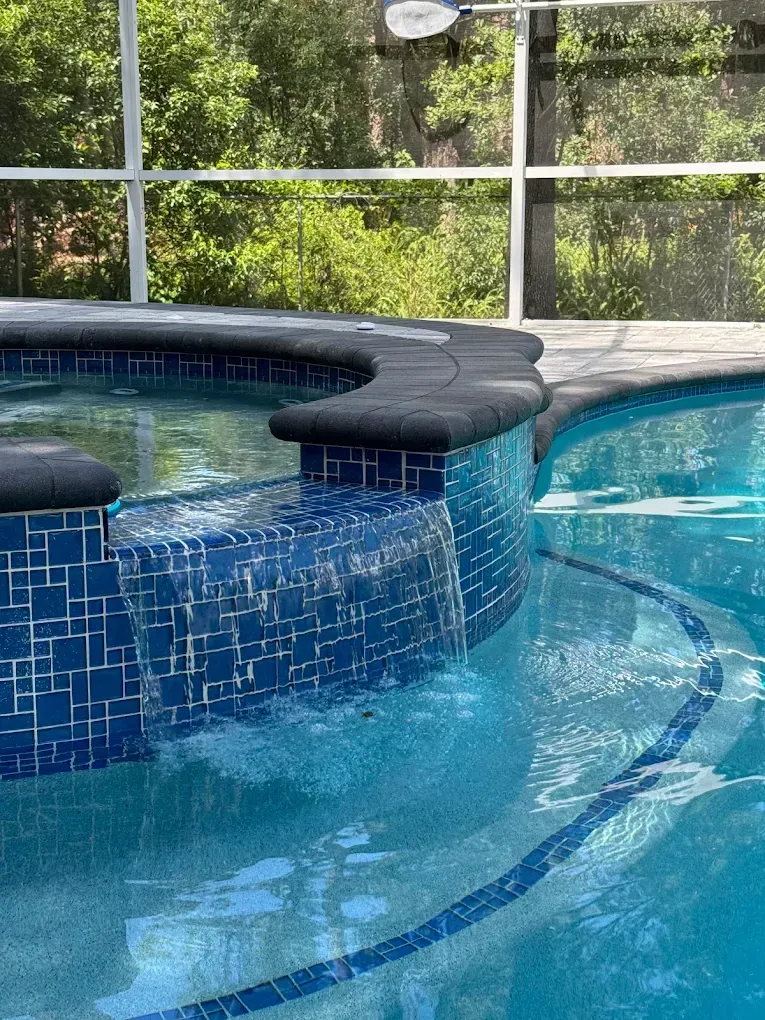 Pool with blue mosaic tiles and a dark ledge, surrounded by a screened enclosure and trees.