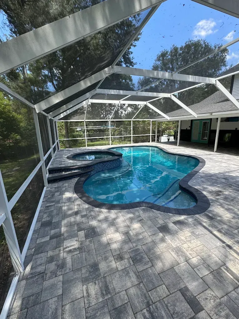 Pool enclosed by a screened lanai with dark pavers and blue water.