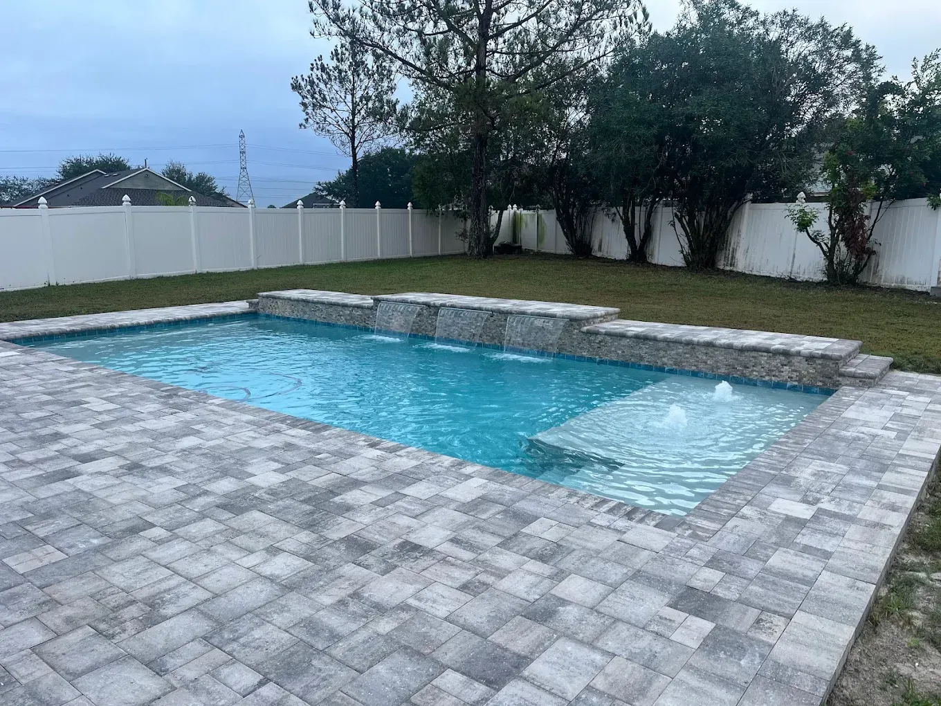 A rectangular pool with waterfall features, surrounded by gray pavers, grass and a white fence.