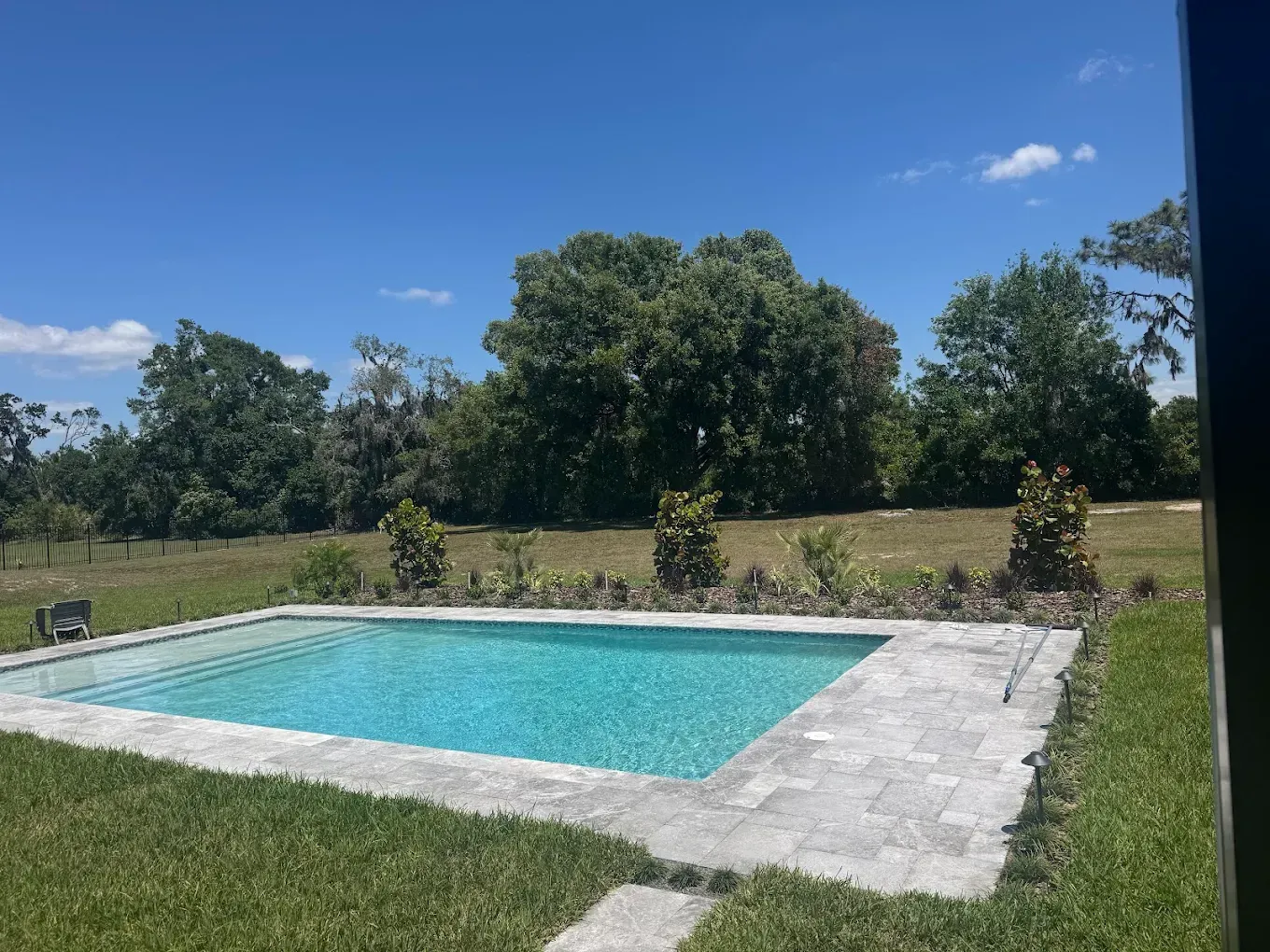 Square swimming pool surrounded by gray stone and green grass under a blue sky, with trees in the background.