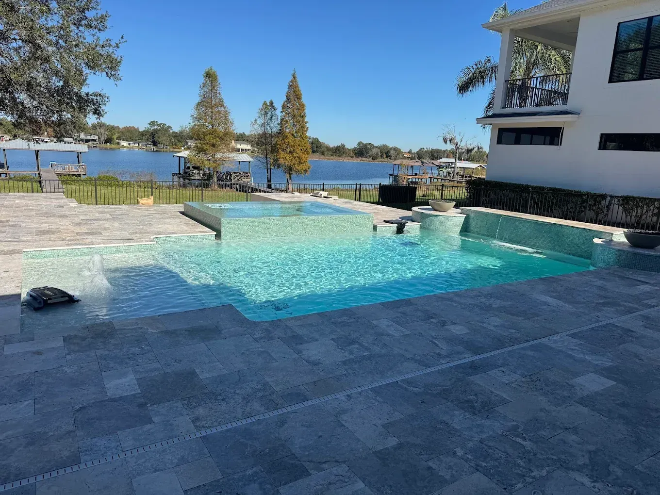 Pool with turquoise water, patio, and lake view from a house on a sunny day.