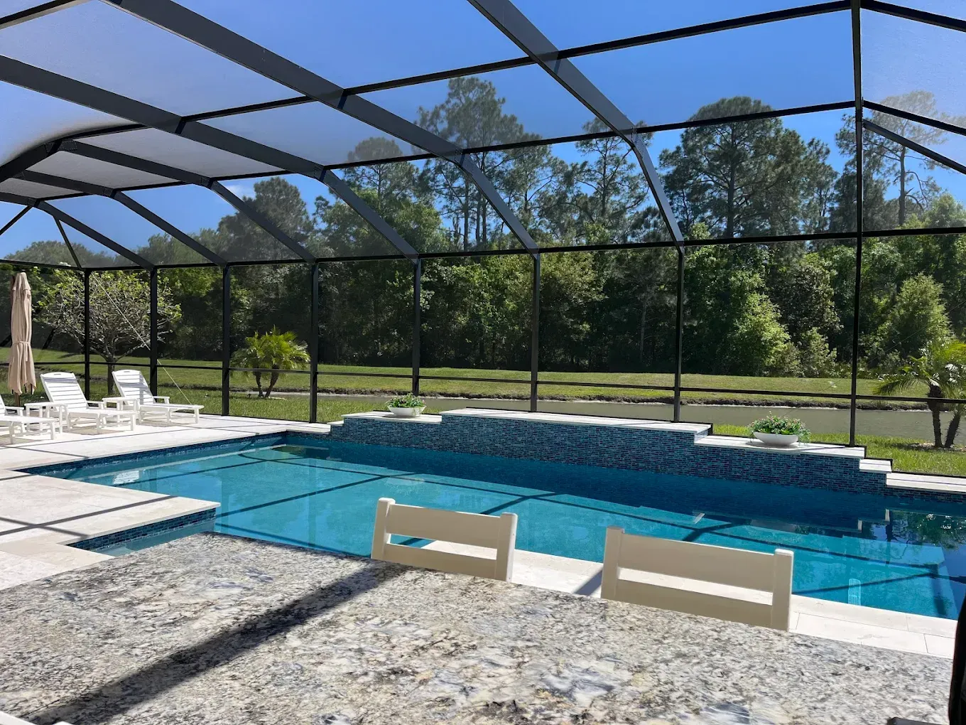 Pool enclosed in screen with trees in the background under a blue sky.