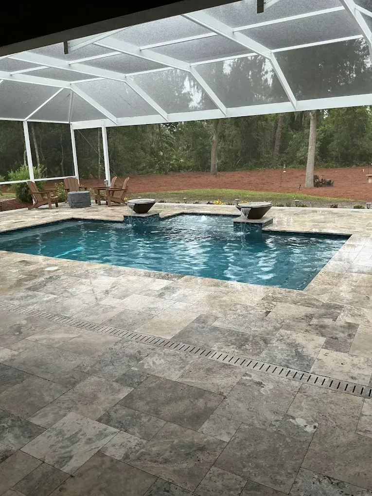Pool with fountains, covered patio, stone deck, and wooded background.