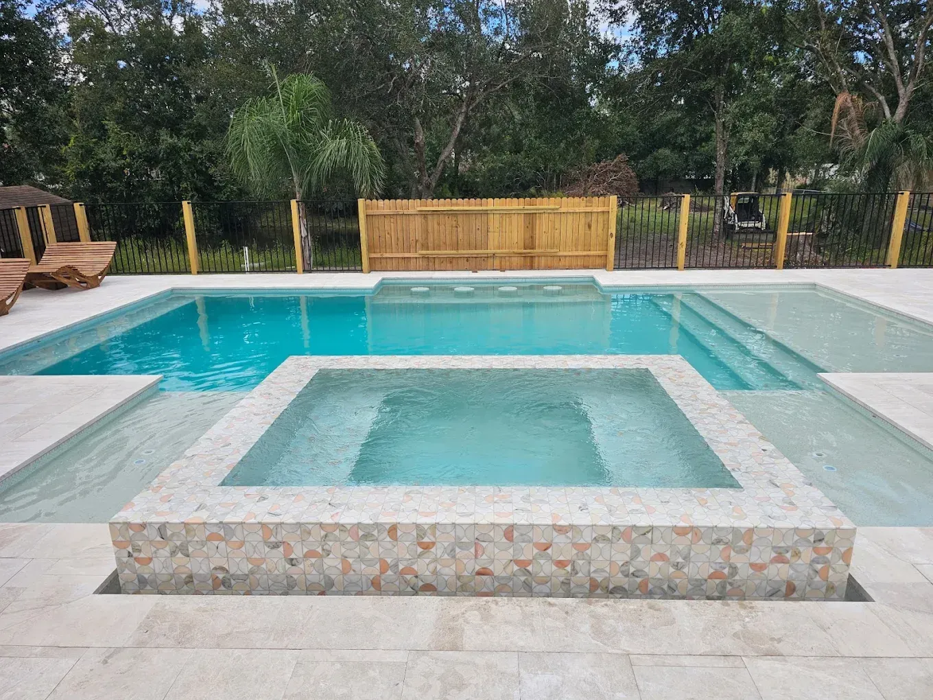 Swimming pool and spa with turquoise water and light-colored tile surround, bordered by a wooden fence and greenery.