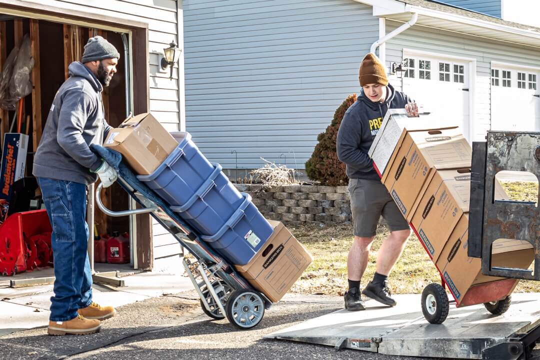 Two movers with stack of boxes on dollies being placed on lift gate to load moving box truck.