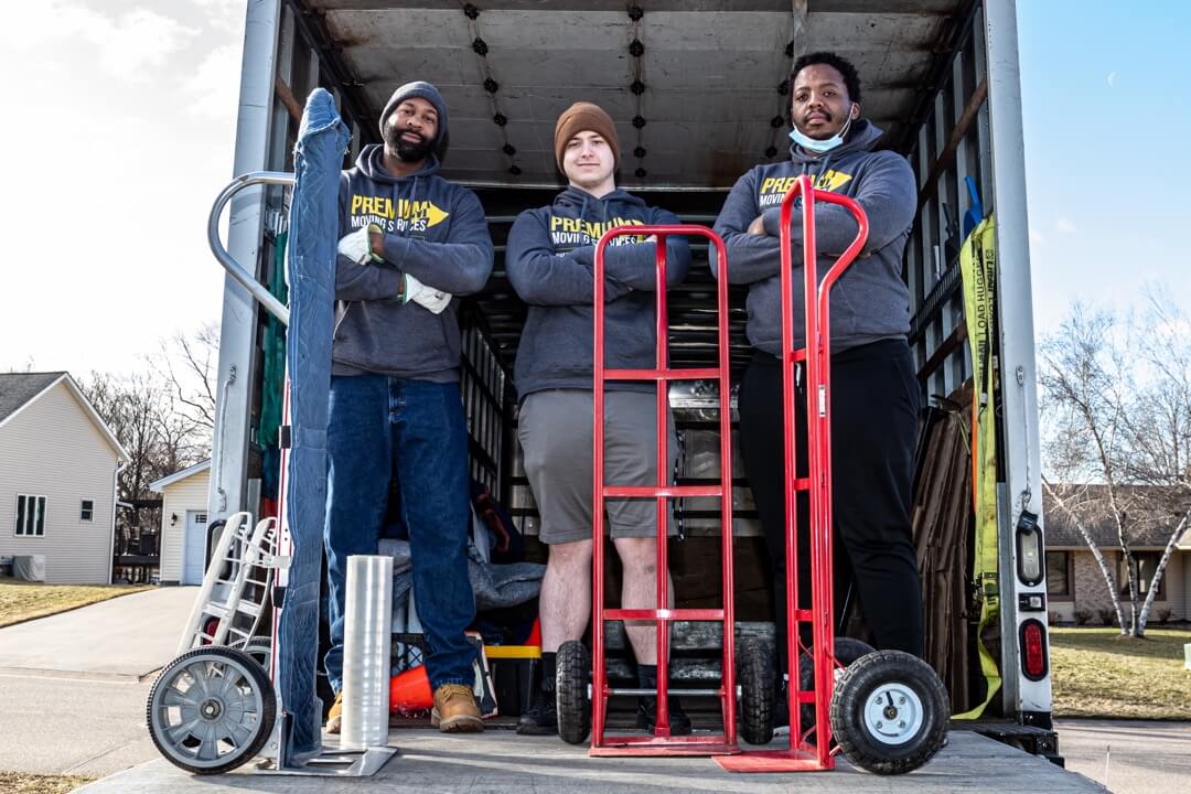 Three professional movers standing on the back of box truck with three two-wheeled dollies.