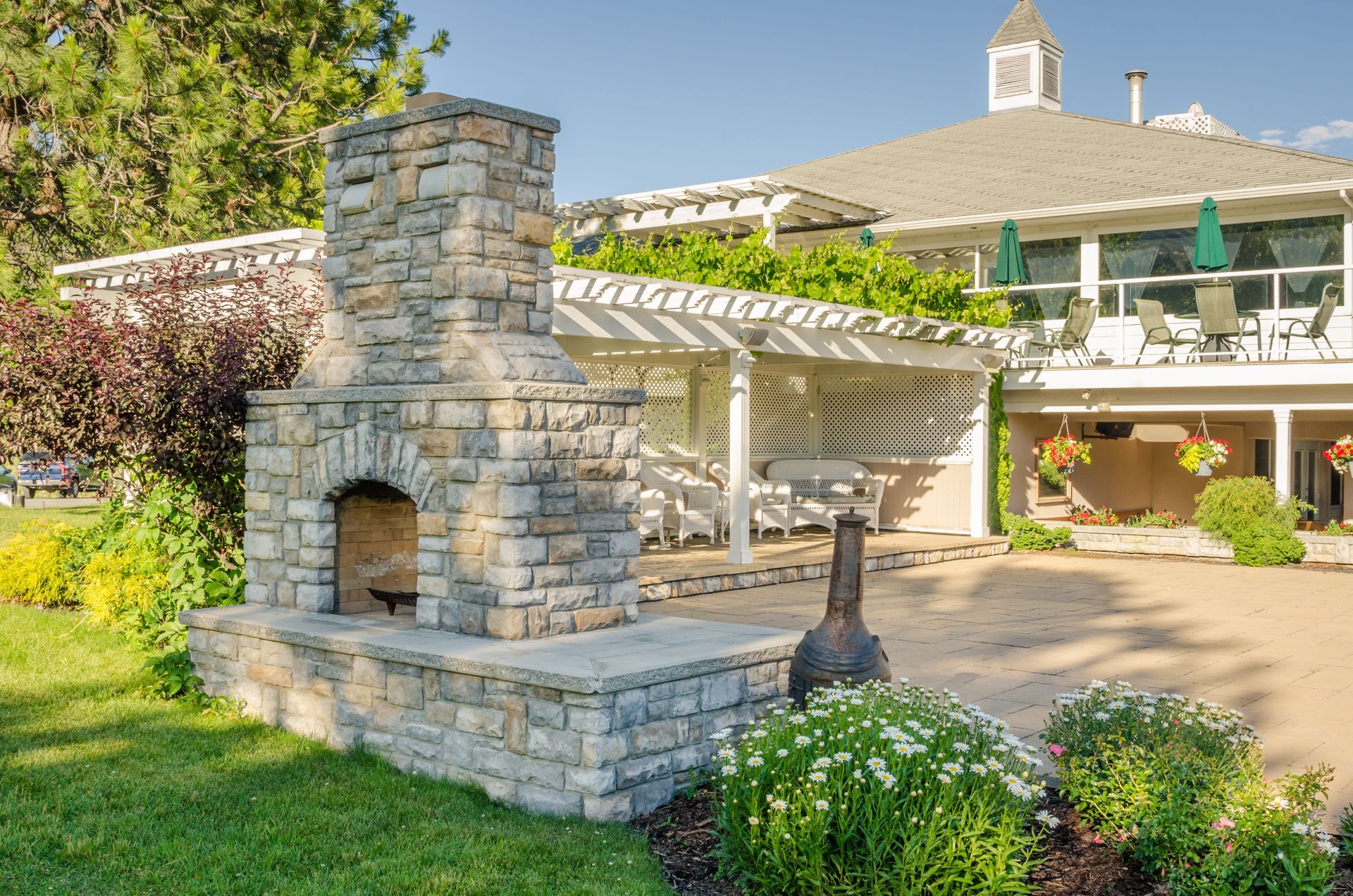 Stone outdoor fireplace with a white pergola and building in the background on a sunny day.
