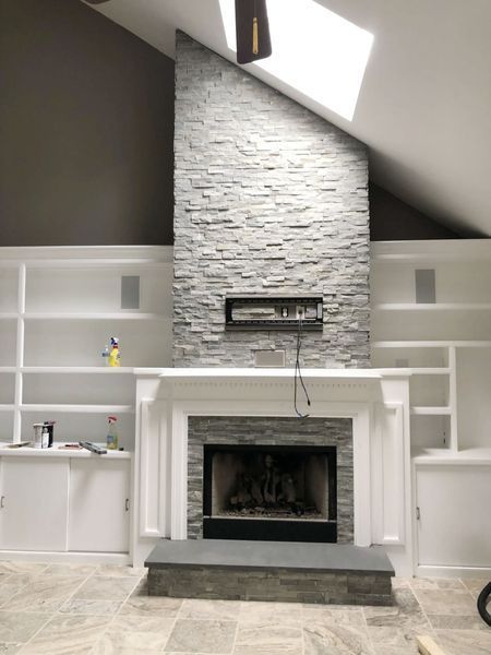 Fireplace with gray stone surround, white built-in shelving, high ceiling, and skylight.