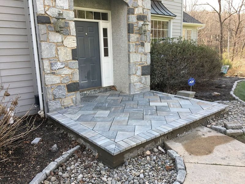 Stone-covered front porch with a gray door, surrounded by stonework, landscaping, and a walkway.