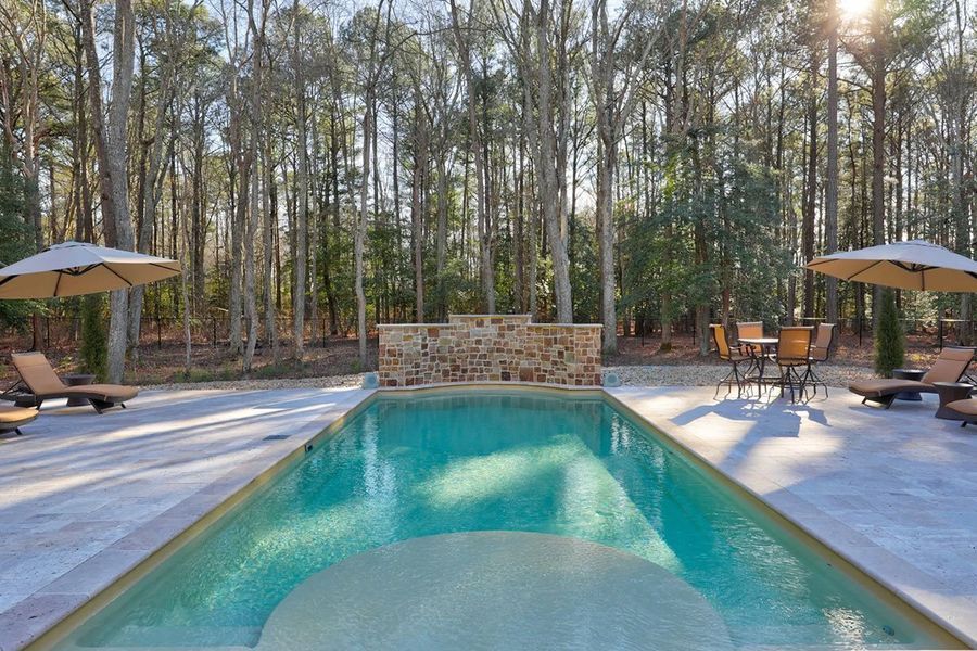 Pool with turquoise water surrounded by a stone patio, lounge chairs, and umbrellas. Forest background.