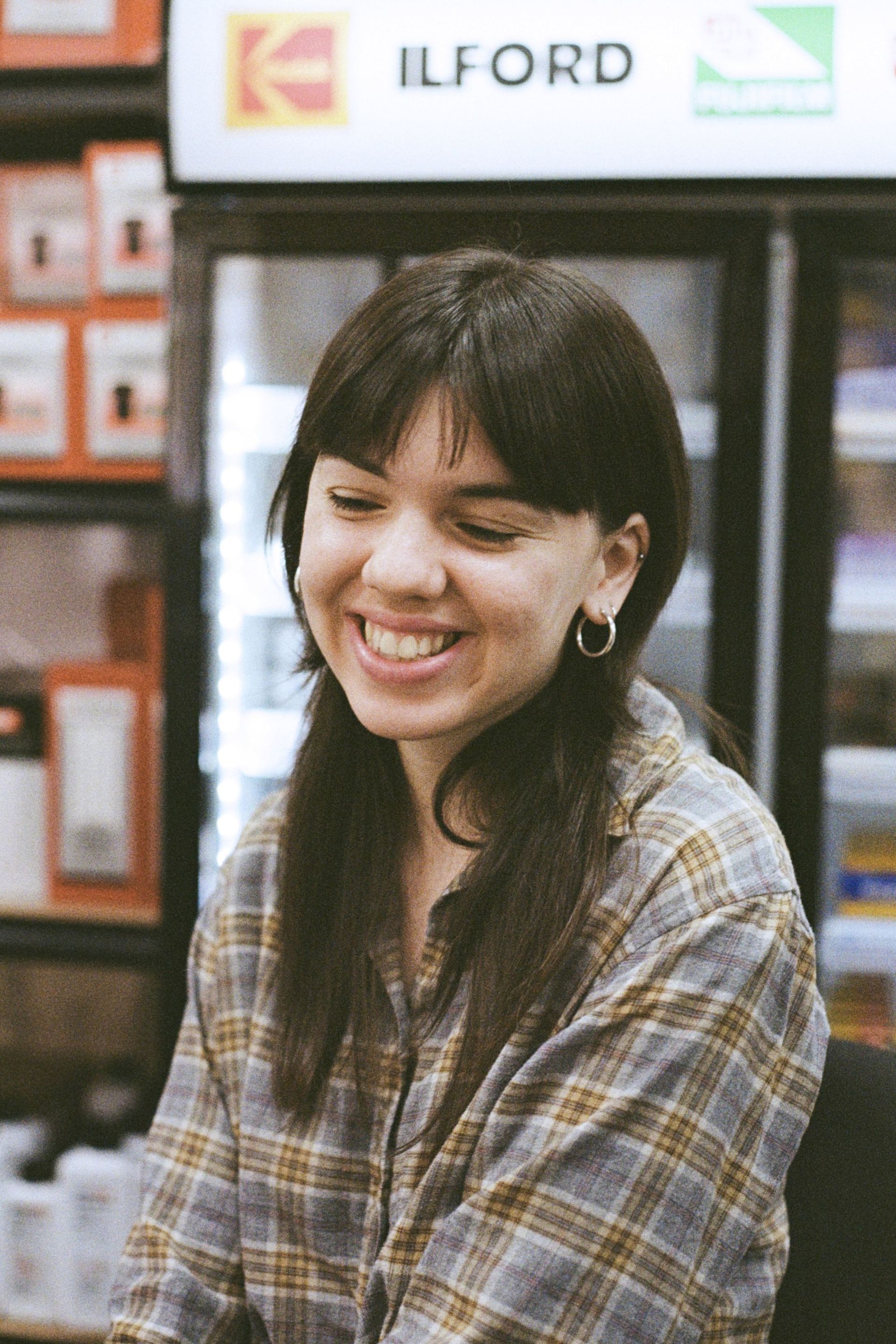 Young woman with dark hair and a plaid shirt smiles broadly inside a store, near a refrigerator.