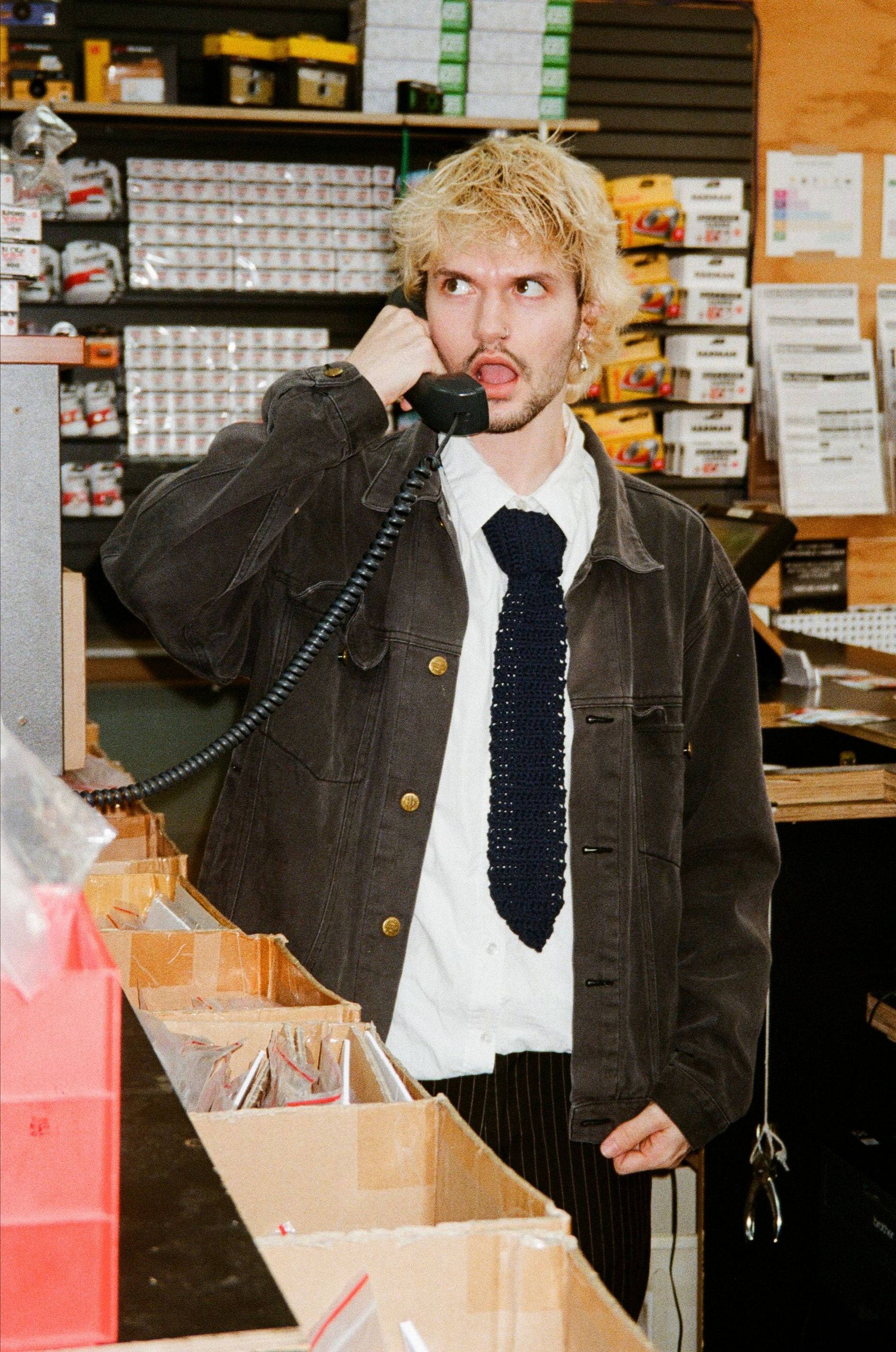 A young man with blonde hair wearing a denim jacket and tie, talking on the phone in a store. He sticks out his tongue.