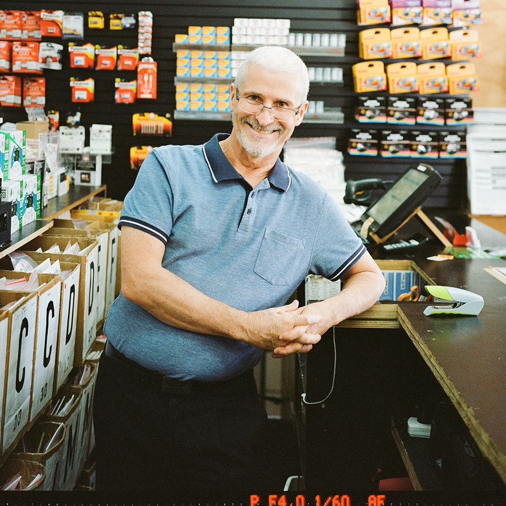 A smiling older man with glasses, wearing a blue polo shirt, leans on a counter in a store.