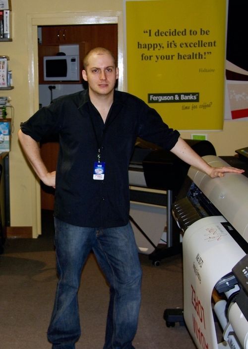 Man in a black shirt and jeans, standing next to a large printer, smiling. A yellow sign is behind him.