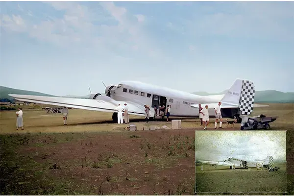 A vintage silver airplane on a grassy field with people boarding. Mountains are in the background under a blue sky.
